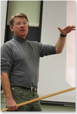 a man stands near a white board looking at an unpictured audience. He is holding a wood pointer in his right hand and gesturing with his left hand.