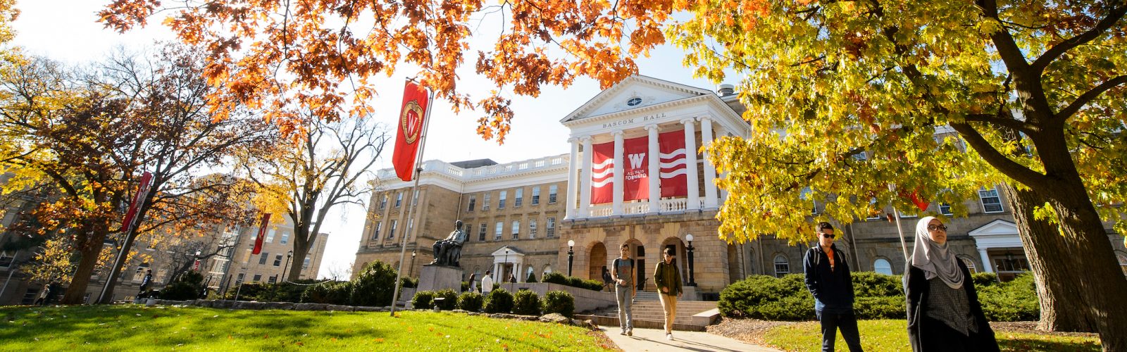 students walking on a path in front of a campus building and surrounded by fall-colored trees