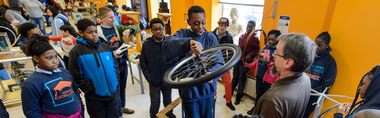 several young students watch in the physics museum as one student holds a spinning bike wheel while standing on a rotatable platform