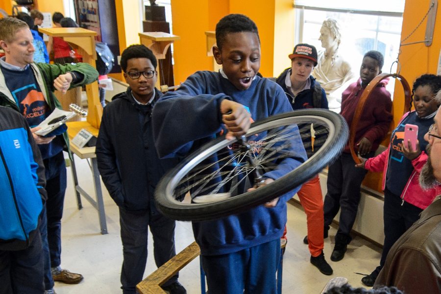 several young students watch in the physics museum as one student holds a spinning bike wheel while standing on a rotatable platform