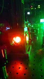 the apparatus hosting the rare earth metal doped crystal glows orange, and the background of the photo has a green fluorescence hue from the input laser