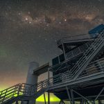 a green, neon-like aurora shines in the bottom of this photo just above the horizon, with the IceCube research station in the foreground