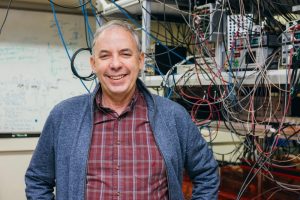 profile photo of Mark Saffman, posing in his lab with lots of wires and equipment