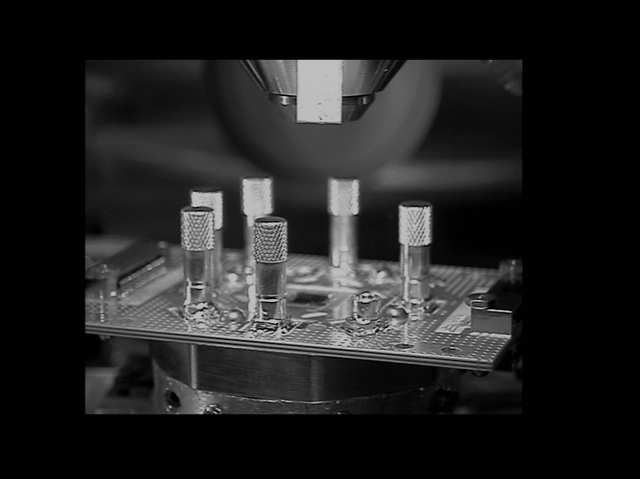 the tip of a scanning electron microscope is poised over a setup with metal clips pointing out