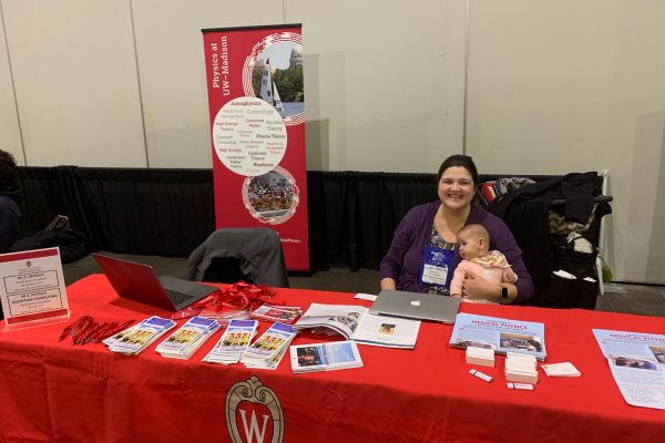 a woman professor holding her baby sits at a table covered in UW physics informational handouts