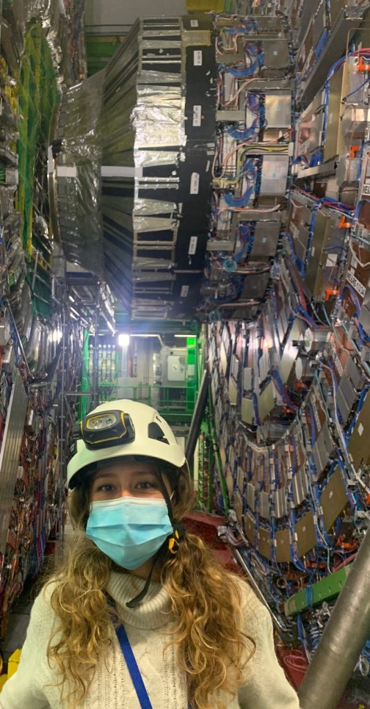 a woman in a helmet wearing a disposable facemask stands in front of lots of metal hardware and wires