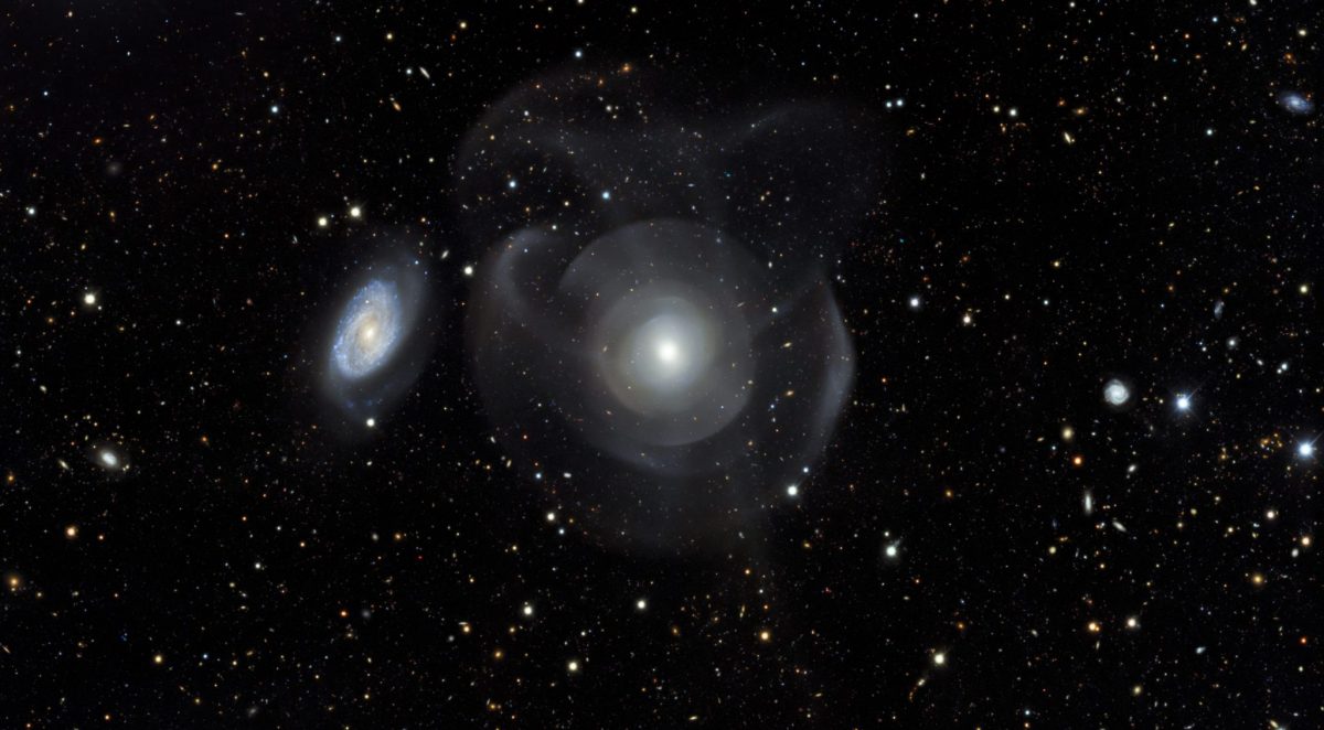 a mostly-black photo of space with white and off-white dots of stars, a small galaxy halo in the left middle, and a larger aura-like glow in the center