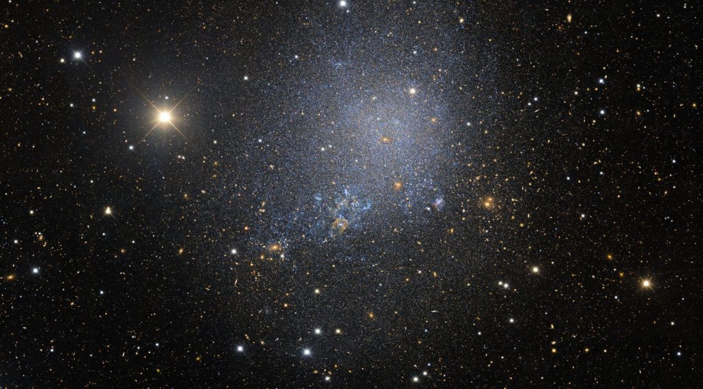 a mostly-black photo of space with white and off-white and blue dots of stars, more concentrated in the middle of the photo and representing the irregular dwarf galaxy