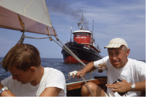 two men on a sailboat with a larger boat behind them