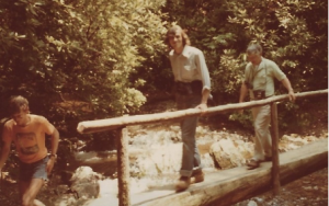 a dated photo showing three men on a tree trunk bridge traversing a river