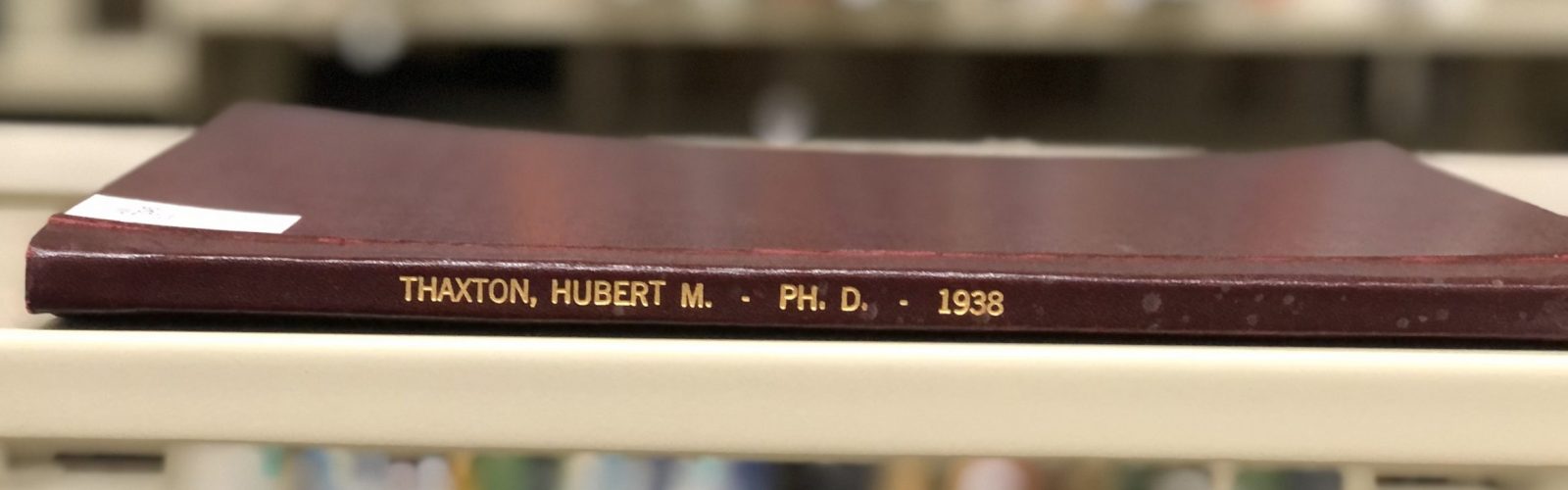 Thaxton's thesis is pictured in the foreground with books blurred in the background behind it