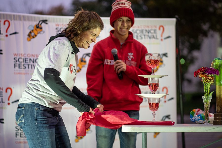 a woman and man on a stage. Man holds microphone, woman is leaning over a table , about to pull a tablecloth on top of which three filled glasses are stacked