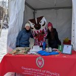 two people stand with Bucky Badger between them, and all three are behind a table that is covered with a red tablecloth that has the department of physics logo on it. They are outside on a frozen lake on a cold day.