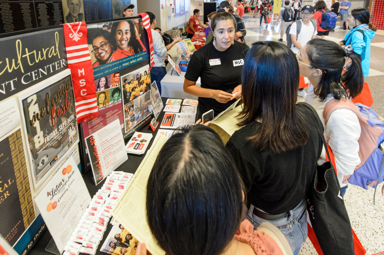 Four people stand in front of a table that holds a poster board and flyers containing information about the Multicultural Student Center. They are inside a campus building at the Fall 2018 Student Organization Fair.