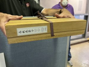 a person, with essentialy just their hands visible, holds a wooden box that is wrapped with a leather tie and has Korean text on the side