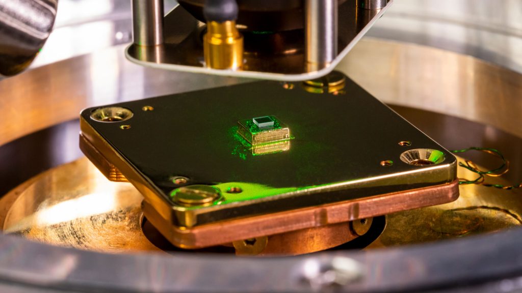 a small square chip sits on a metallic microscope stand with green laser light bouncing off of it in places