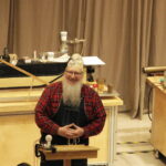 a man stands behind a lectern with physics gadgets behind him. he is wearing a costume that centers around the theme of time.