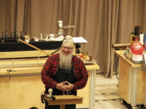 a man stands behind a lectern with physics gadgets behind him. he is wearing a costume that centers around the theme of time.