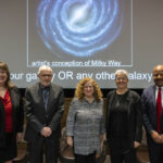 a group of 5 people stand in front of a screen that is showing an artist's rendering of the Milky Way