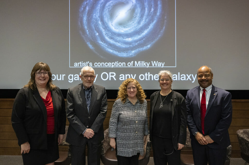 a group of 5 people stand in front of a screen that is showing an artist's rendering of the Milky Way