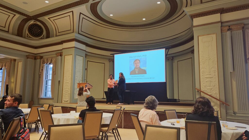 a zoomed out photo shows a man receiving an award from a woman on an elevate stage, with a screen behind them showing his photo, name, and award won