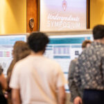 the focus of this image is on the background, with research posters tacked to boards and a sign above that says "undergraduate symposium." The blurry foreground shows attendees of the symposium looking at a poster. their backs are all to the camera