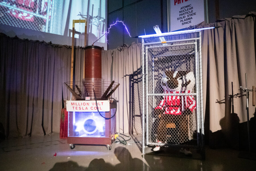 The university mascot, Bucky Badger, sits in a metal cage with a spark of electricity visibly connecting to the cage from a Tesla coil apparatus to the left of the cage