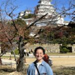 a woman stands in front of a tree with a Japanese castle in the near background
