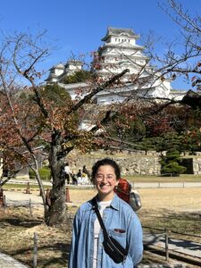 a woman stands in front of a tree with a Japanese castle in the near background
