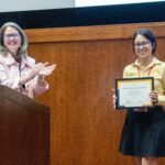 two women stand on a stage. the one on the left is behind a podium clapping, the one on the right is holding a frame with a certificate in it