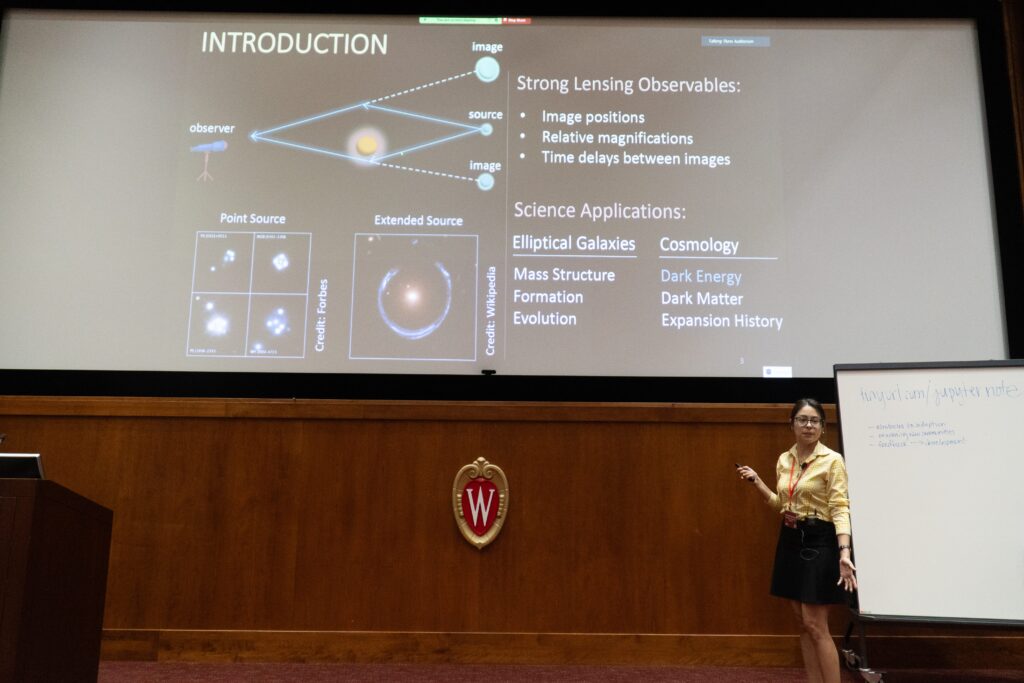 a woman stands in front of a screen with a research slide on the screen, she faces the audience and is gesturing with her hands. 