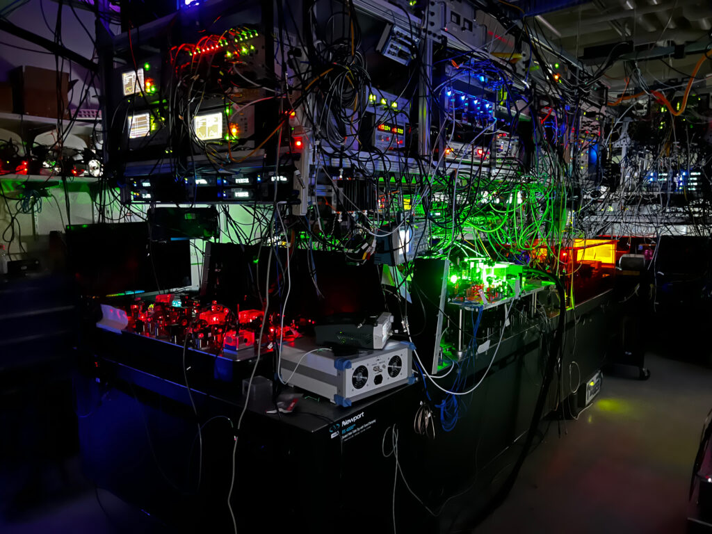 a photograph of a room with the lights off, but the bulk of the image is taken up by a large piece of complicated equipment with many different colored laser lights visible, illuminating the shape of the equipment