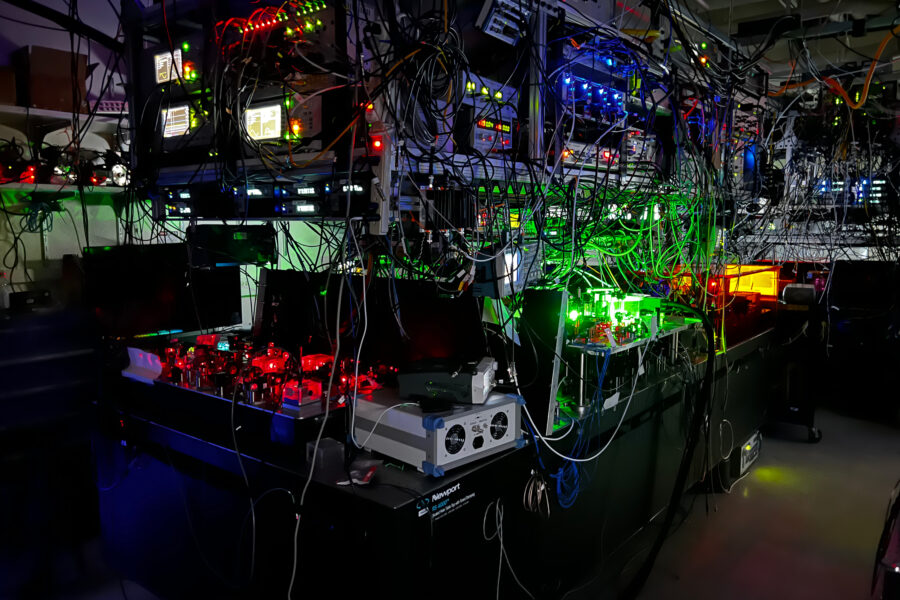 a photograph of a room with the lights off, but the bulk of the image is taken up by a large piece of complicated equipment with many different colored laser lights visible, illuminating the shape of the equipment
