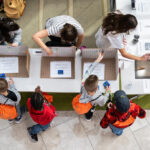 a bird's eye view of a science fair table, with three presenters showing 5 visitors a demo