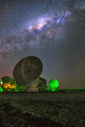 a series of dish antenna telescopes is gently illuminated under the night sky with the milky way galaxy visible above