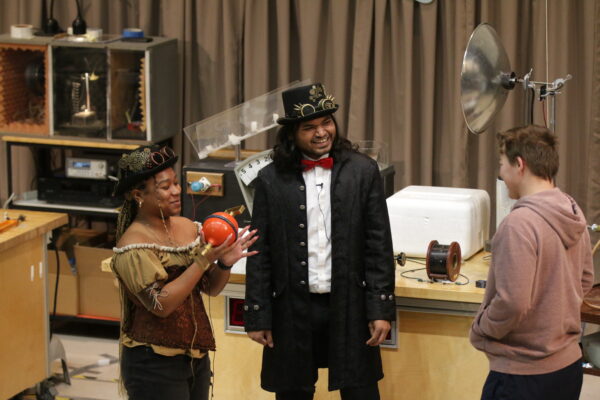 a woman is on the left, holding two orange half spheres. In the middle is a man. Both are wearing steam punk outfits. An older child is on the right, facing the other two.