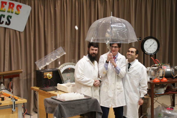 three men stand under an umbrella as small white objects fly by in the air (the objects are plastic film cannister lids, but they are too hard to discern with how fast they are moving)