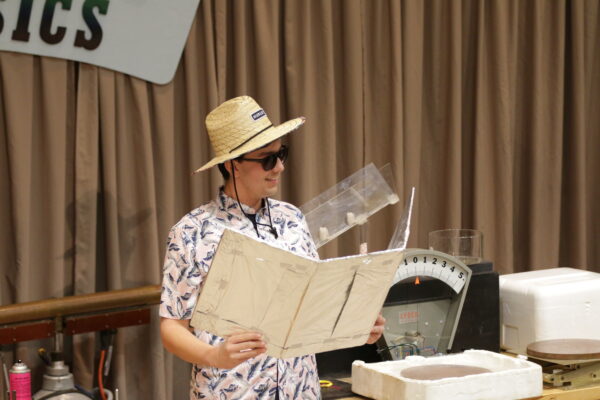 a man wearing a sun hat, sunglasses, and a floral-print shirt holds a folding board, meant to reflect the sun, in front of him