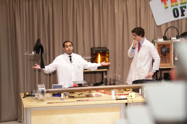 two men wear lab coats and stand behind a large block table.