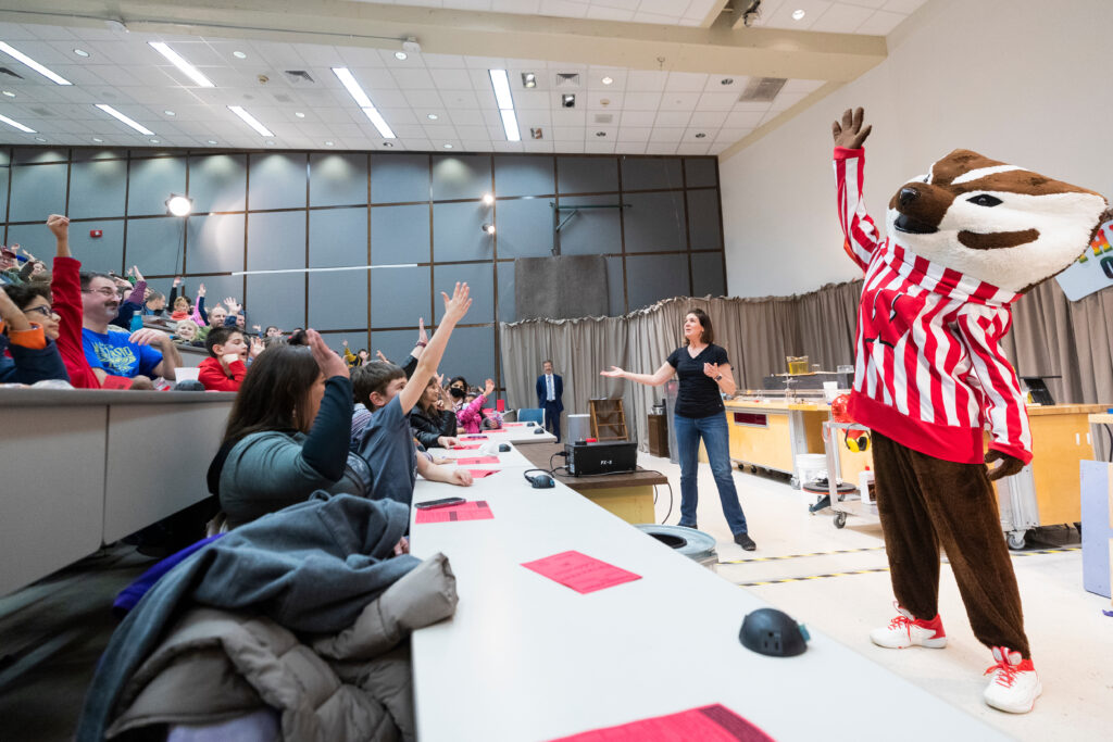a woman on the left of a stage and Bucky Badger on the right look toward the audience, pictured to the left of the photo, to recruit volunteers