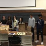 a group of 7 high school students stand behind a table holding cardboard and wooden prototypes of the demo they developed