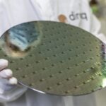hands from an unidentified individual hold a metal pancake-like object with some tech-y designed on it. The object is roughly a foot in diameter.