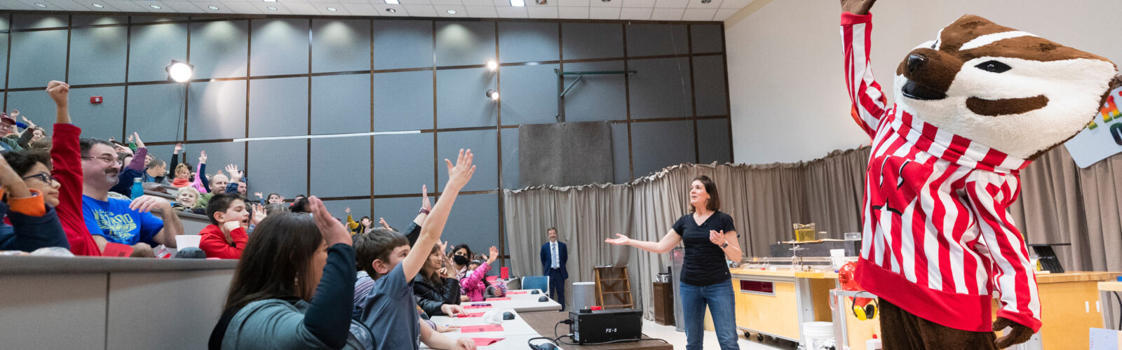 bucky badger raises his hand in front of an audience of enthusiastic kids at a Wonders of Physics show