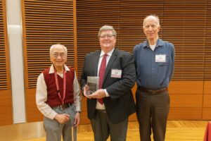 three men stand, one is holding a glass plaque