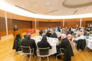 a woman stands at a podium while people seated at a table look on