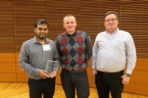 three men stand, one is holding a plaque