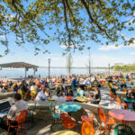 a crowded terrace with tables and lots of people sitting at them on the shore of a lake