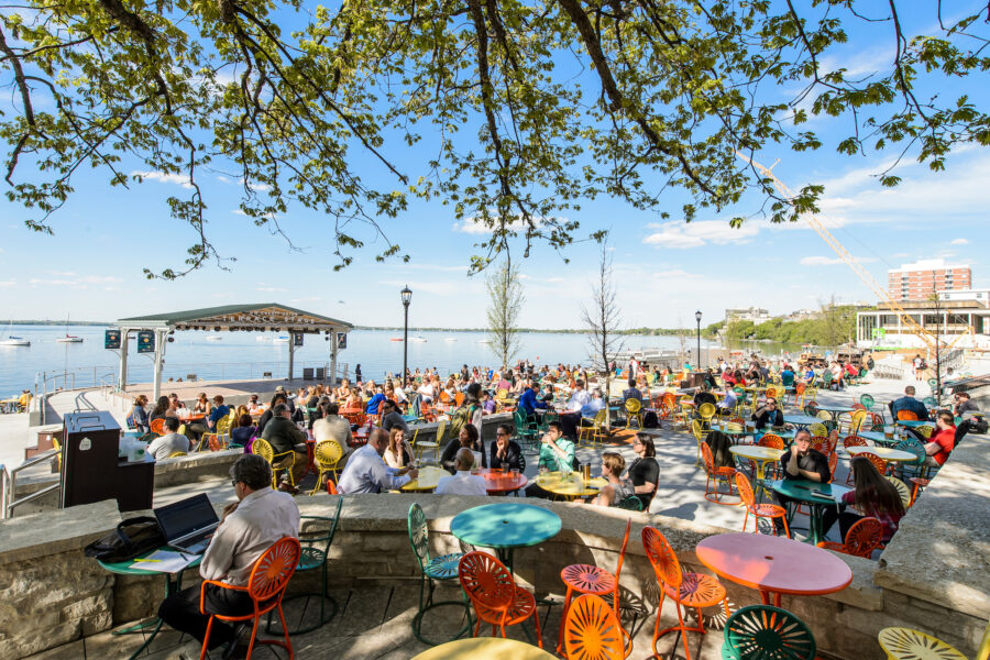 a crowded terrace with tables and lots of people sitting at them on the shore of a lake