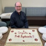 a man sits behind a cake that says "Congrats Prof. Rzchowski"