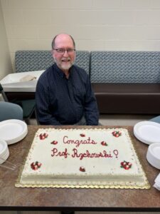a man sits behind a cake that says "Congrats Prof. Rzchowski"
